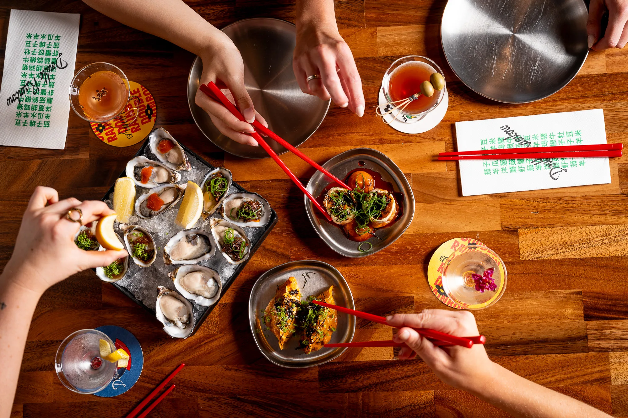 Hands use red chopsticks to pick up food from small plates of oysters and other dishes on a wooden table.