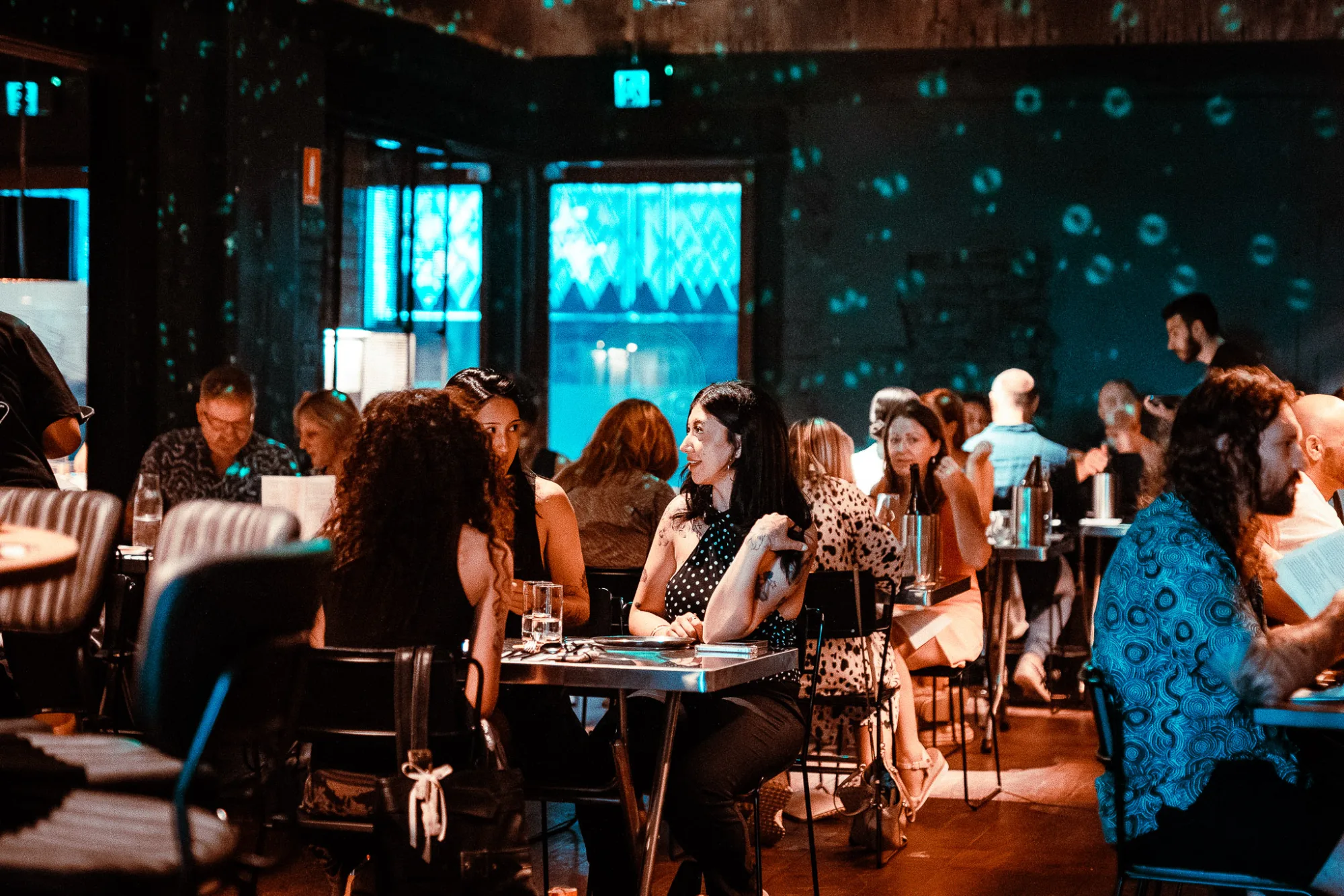 People dining and socializing at tables in a dimly lit restaurant with blue light projections on the wall.