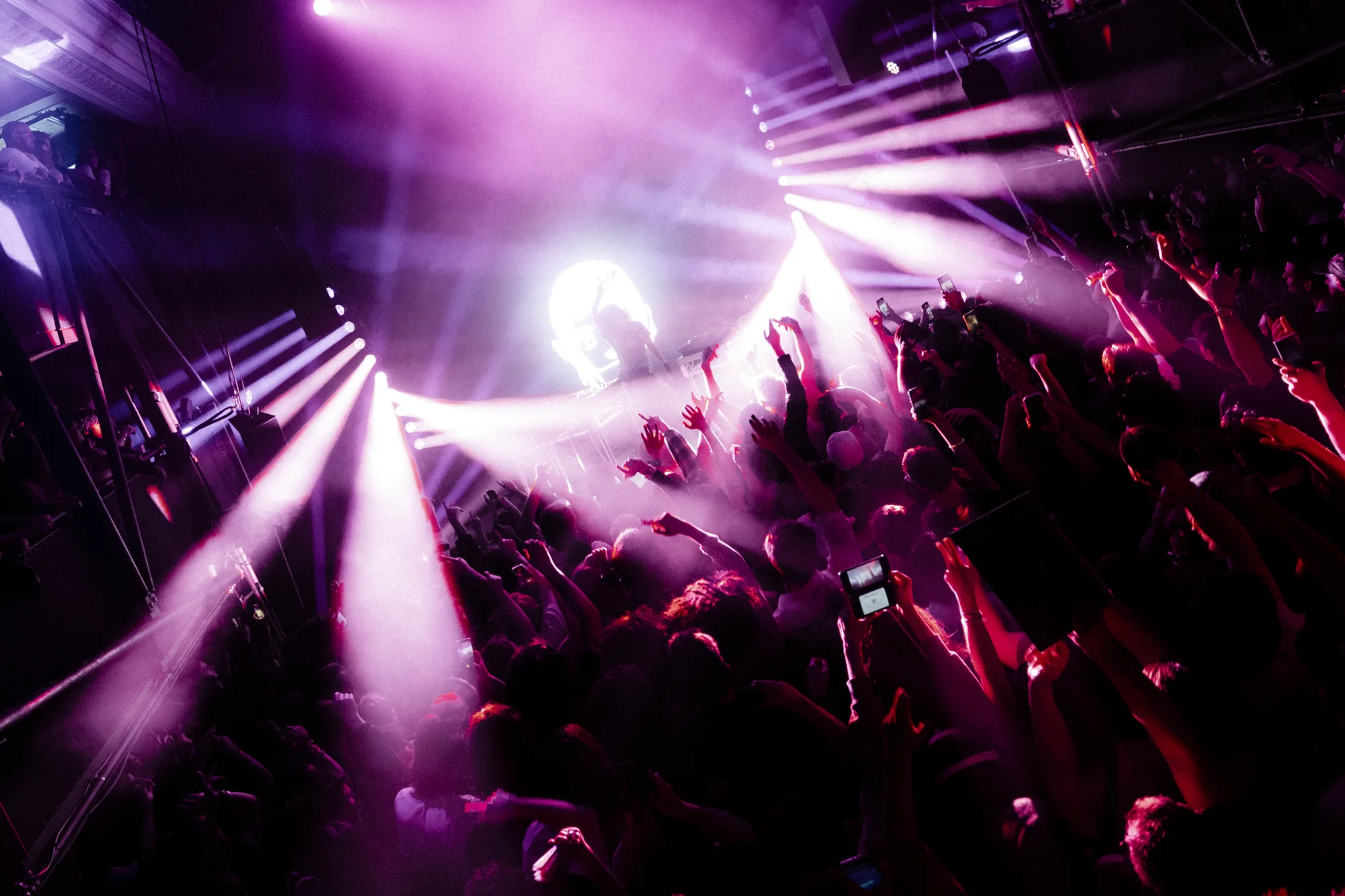 A crowd of people with their hands in the air at a concert, illuminated by purple stage lights.