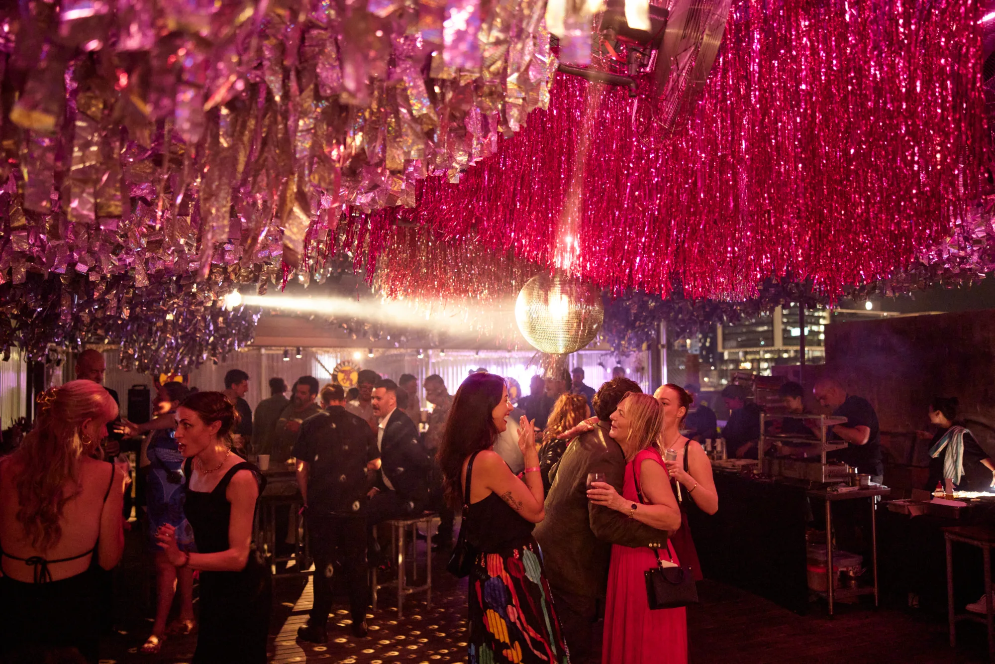 A rooftop bar decorated with pink and silver tinsel and a disco ball, filled with people socializing.