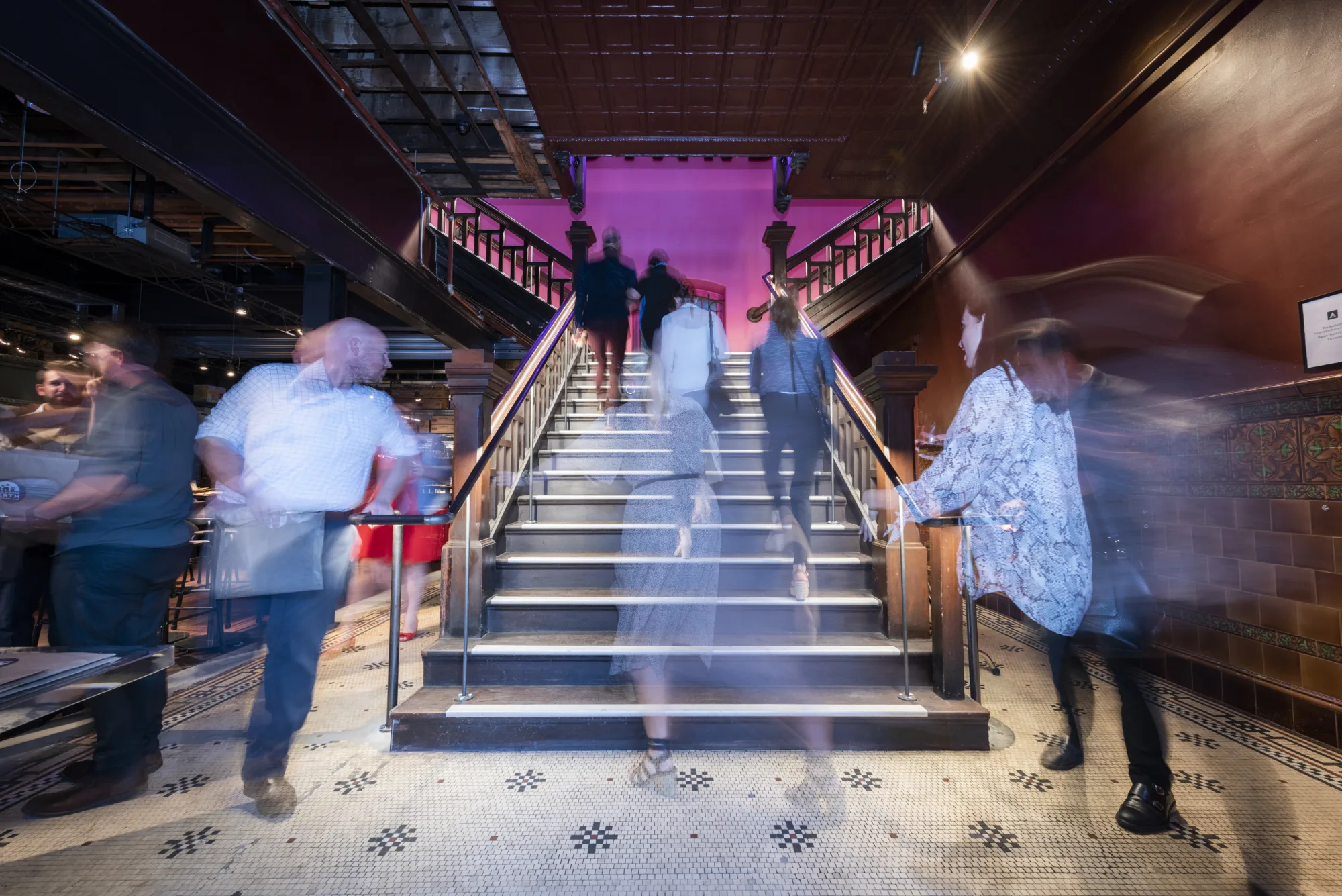 People ascend and descend a grand staircase with pink lighting and ornate railings.