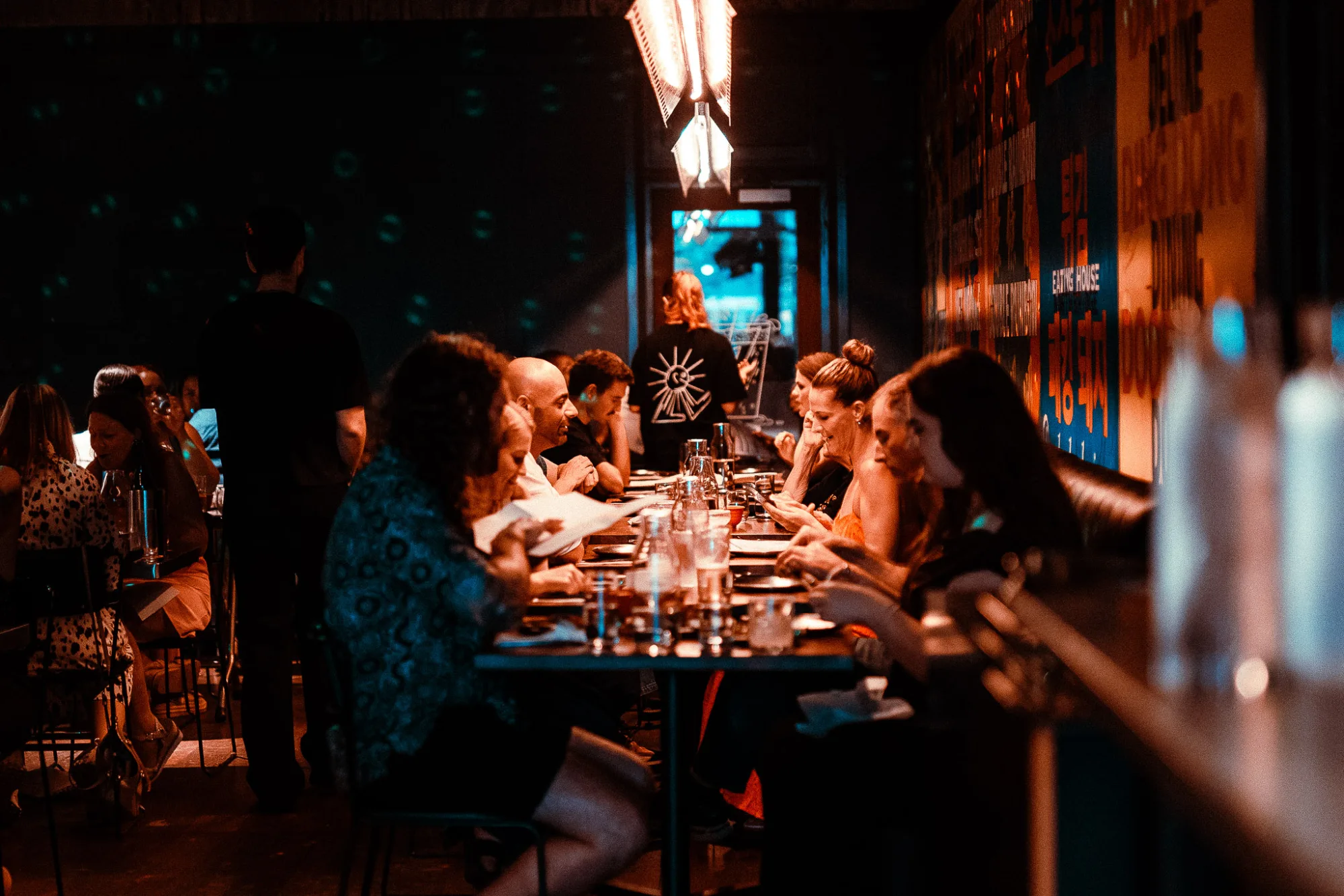 A group of people dine at a long table in a dimly lit restaurant with a unique light fixture overhead.