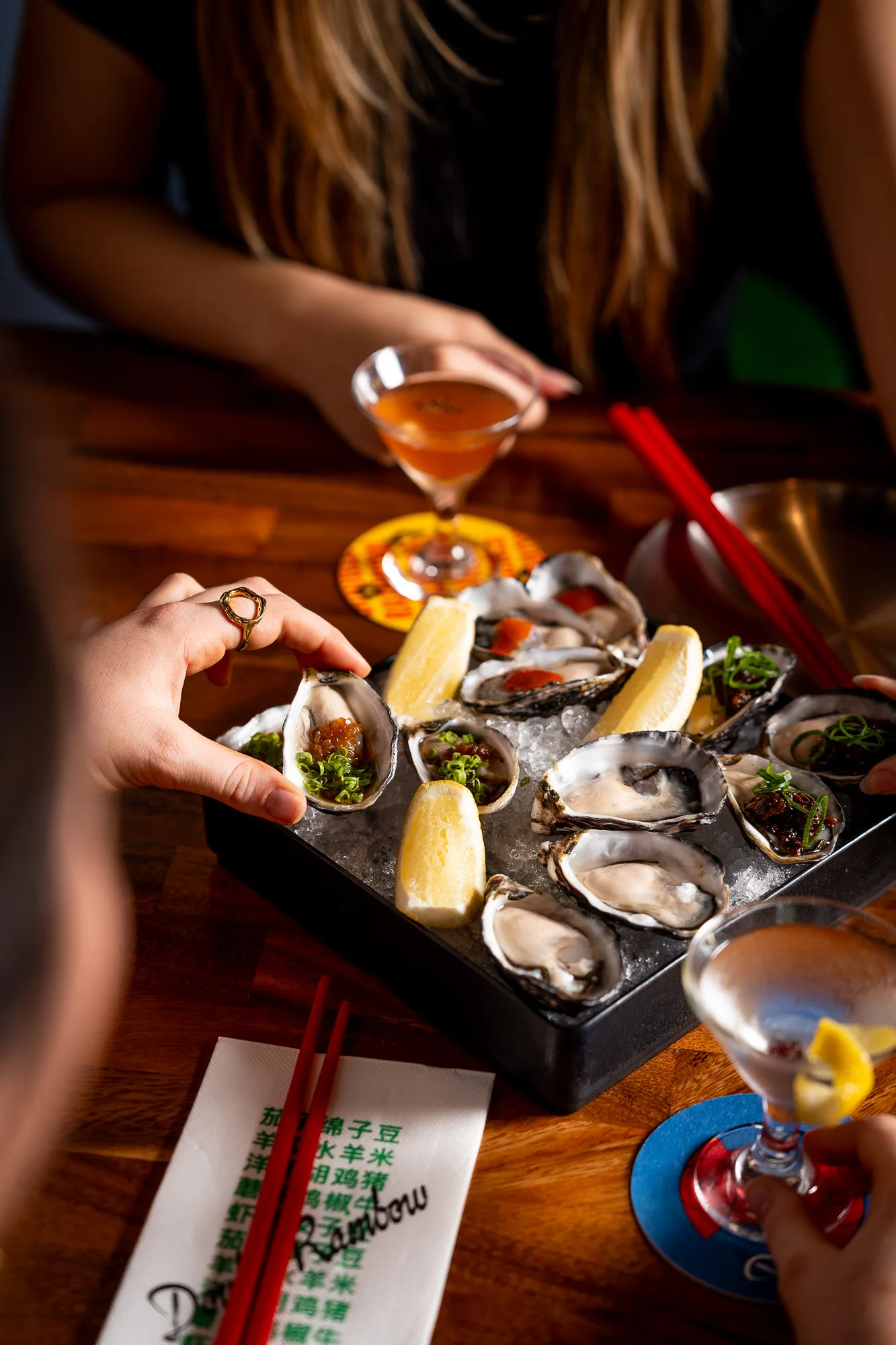 A hand picks up a fresh oyster from a platter of oysters on ice, served with lemon wedges and garnishes.
